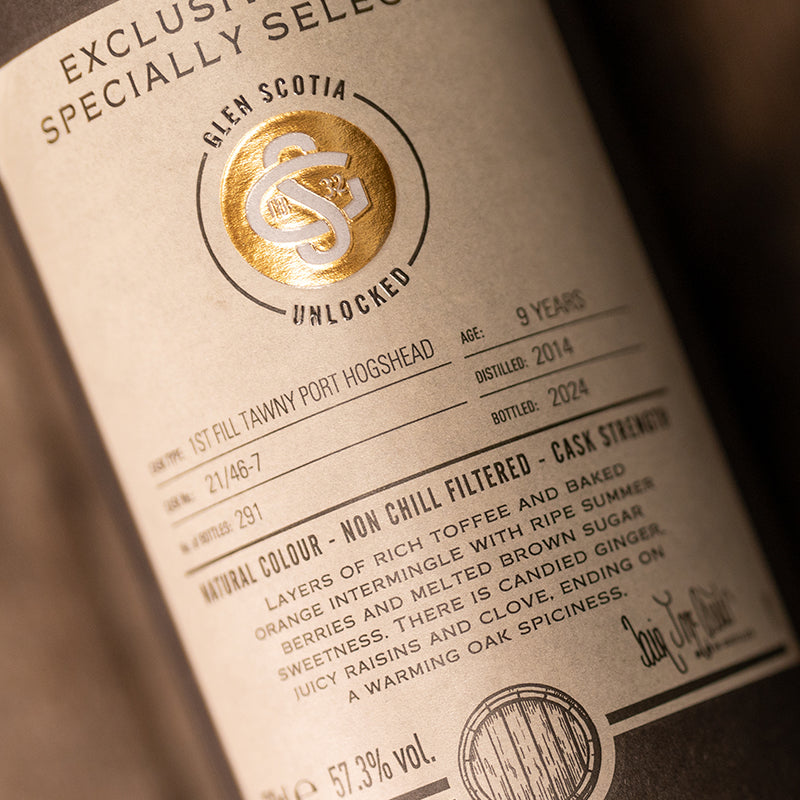 A close-up of an Unlocked #1 9 Year Old Single Malt bottle label from Glen Scotia highlights an exclusive cask. It describes a whiskey aged for 9 years in 1st fill Tawny Port casks, bottled in 2024, featuring notes of toffee, orange, and spices. The text prominently includes "Natural Colour" and "NON CHILL FILTERED.