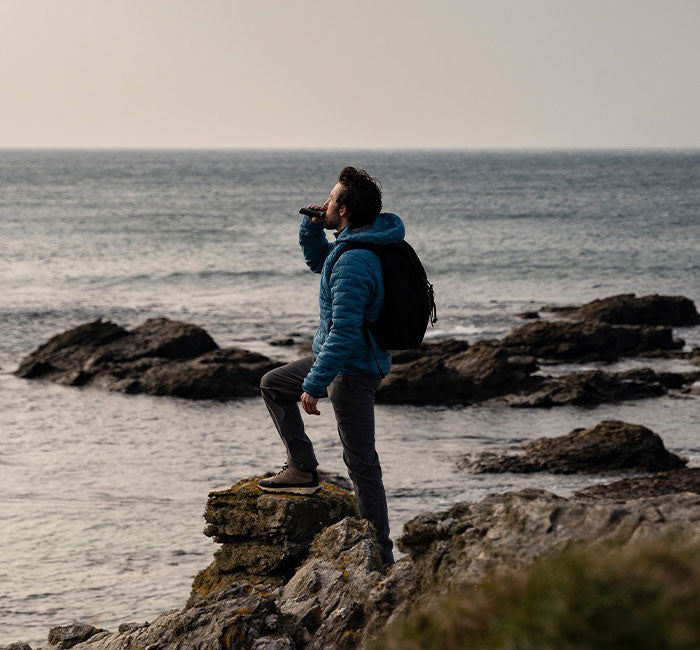 A person wearing a blue jacket and a black backpack stands on a rocky shore, gazing out at the ocean while holding a camera or binoculars to their eyes. The sea stretches out under a cloudy sky in the background.