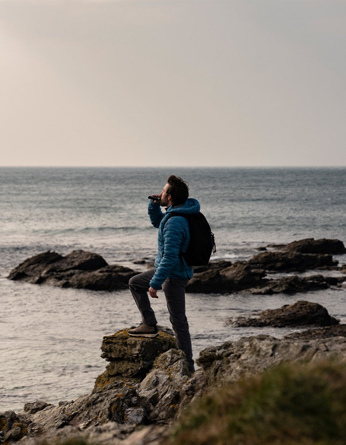 A person wearing a blue jacket and a black backpack stands on a rocky shore, sipping from a bottle while looking out over the ocean. The sky is overcast, and the water is calm with gentle waves.