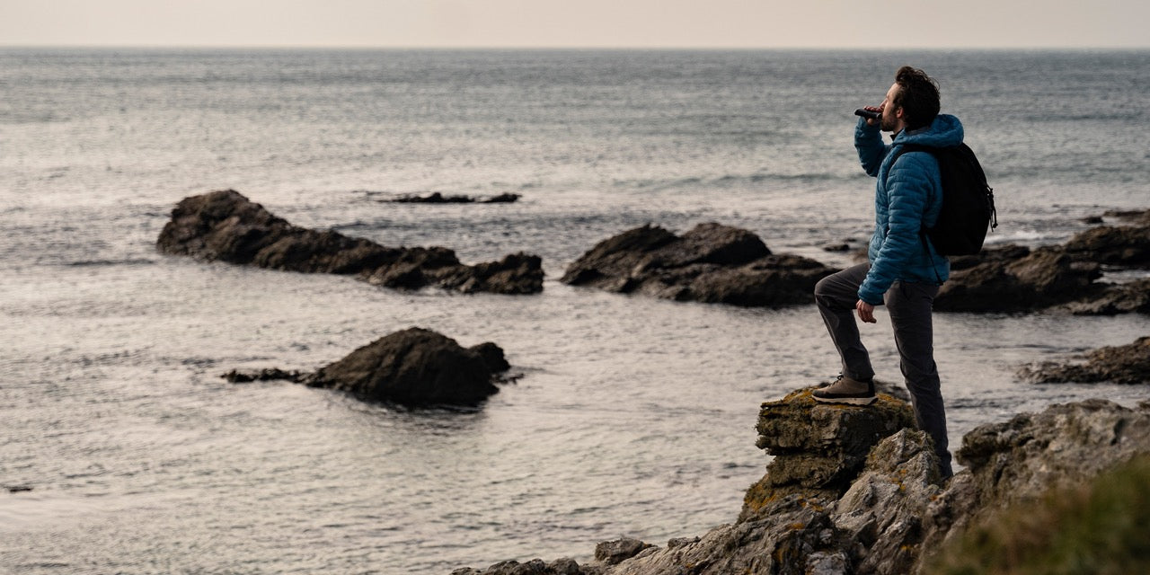 A man in a blue jacket and gray pants stands on a rock near the ocean. He has a backpack and is drinking from a bottle while looking at the water. The sky is overcast, and there are rocky formations and waves in the background.