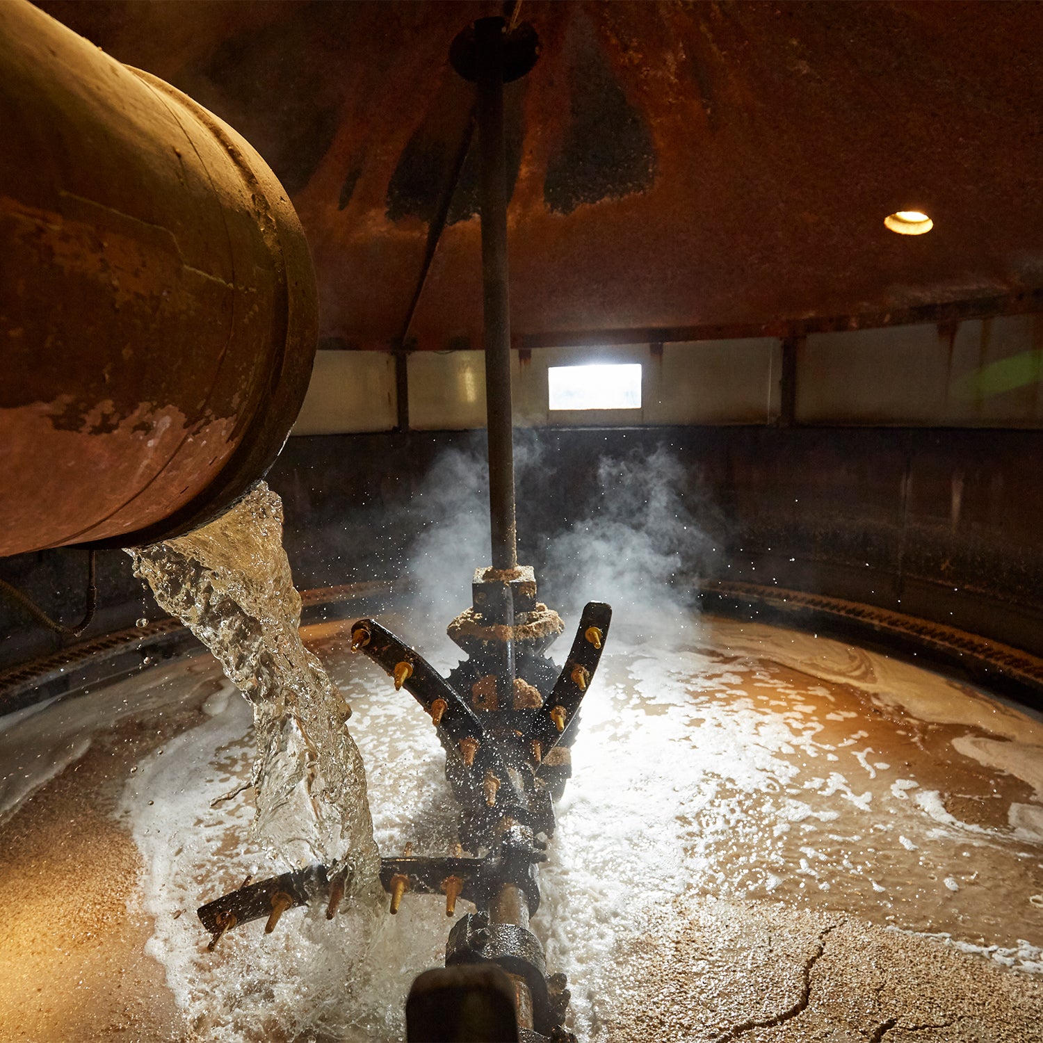 A large brewing vessel with water spraying from multiple nozzles on a pipe, creating steam and splashing on the sides. Warm light illuminates the interior, highlighting the metal surfaces and liquid movement inside the container.