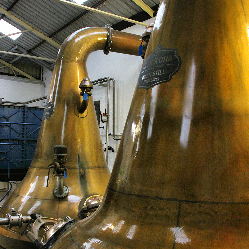 Large copper stills inside a distillery, used for the production of whisky. The stills are shiny and reflective, set against an industrial background with metal pipes and a corrugated roof. An attached plaque indicates the capacity and type of the still.