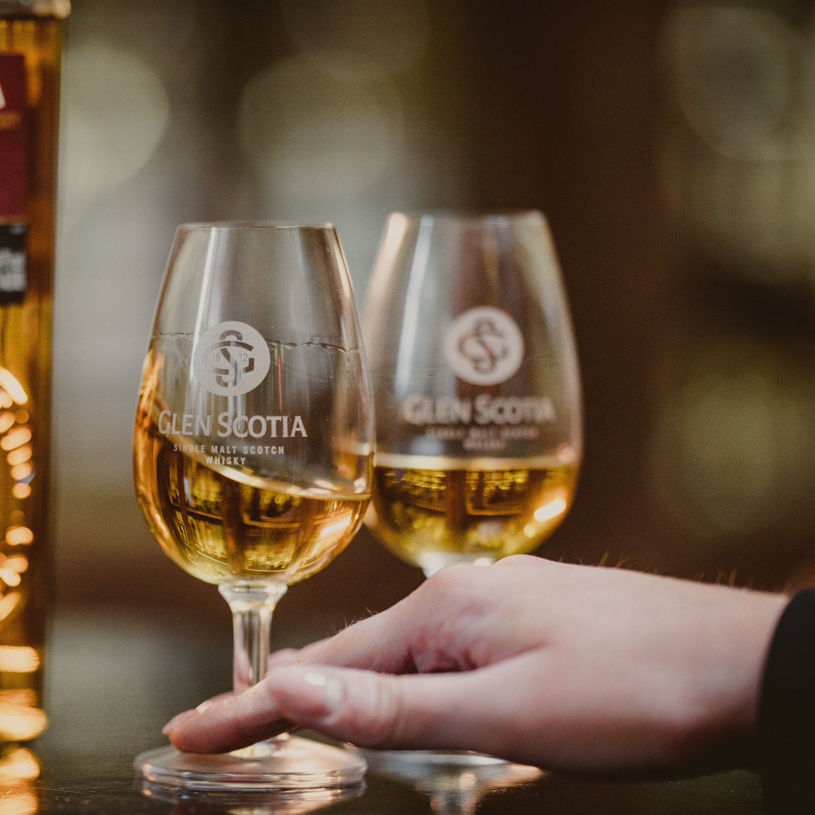 A close-up of a person's hand holding a glass of Glen Scotia single malt Scotch whisky. Another filled glass and a Glen Scotia whisky bottle are in the background. The setting appears to be a dimly lit, warm-toned environment.
