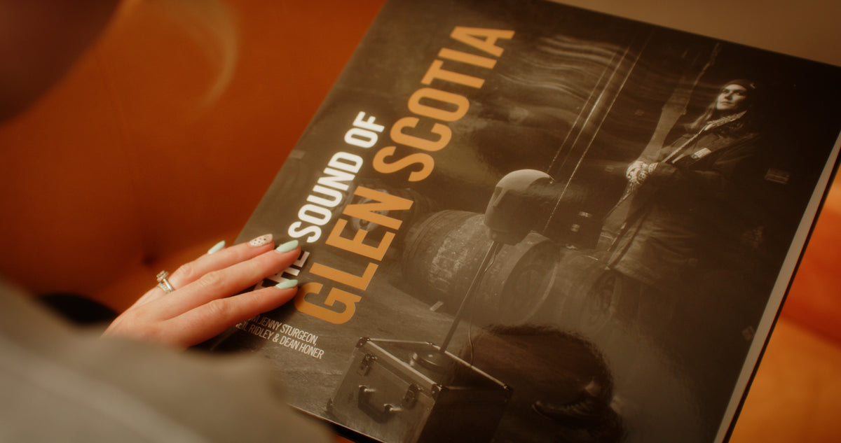 A person holds a vinyl record with the title &quot;THE SOUND OF GLEN SCOTIA&quot; from Glen Scotia written on the cover. The cover features someone in a kilt and traditional Scottish attire, standing next to whisky barrels in Campbeltown.