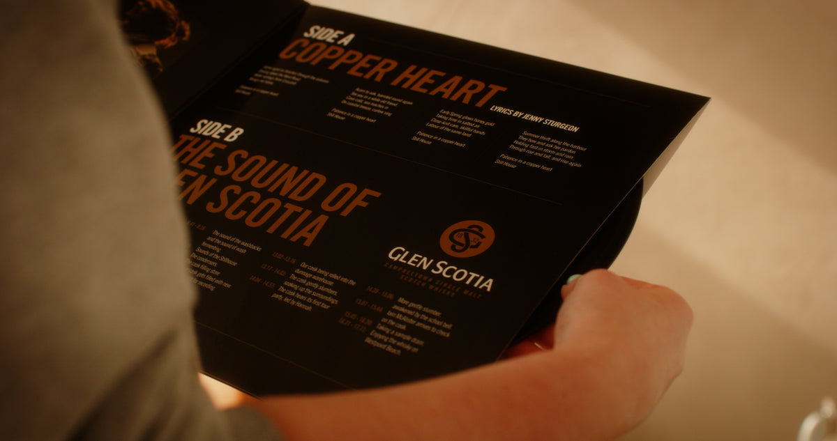 A person holds &quot;The Sound of Glen Scotia,&quot; a black booklet from the brand Glen Scotia. It features two titled sections: &quot;Side A: Copper Heart&quot; and &quot;Side B: The Sound of Glen Scotia.&quot; Each section lists events or items with descriptions. The iconic Glen Scotia logo, synonymous with Campbeltown whisky, is visible in the lower half of the right page.