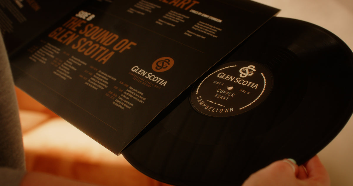 Close-up of a person holding a vinyl record partially pulled out from its sleeve. The sleeve features a design with text about &quot;The Sound of Glen Scotia,&quot; and the label on the record reads &quot;Glen Scotia.&quot; The warm-toned, blurred background evokes the rich heritage of Campbeltown whisky.