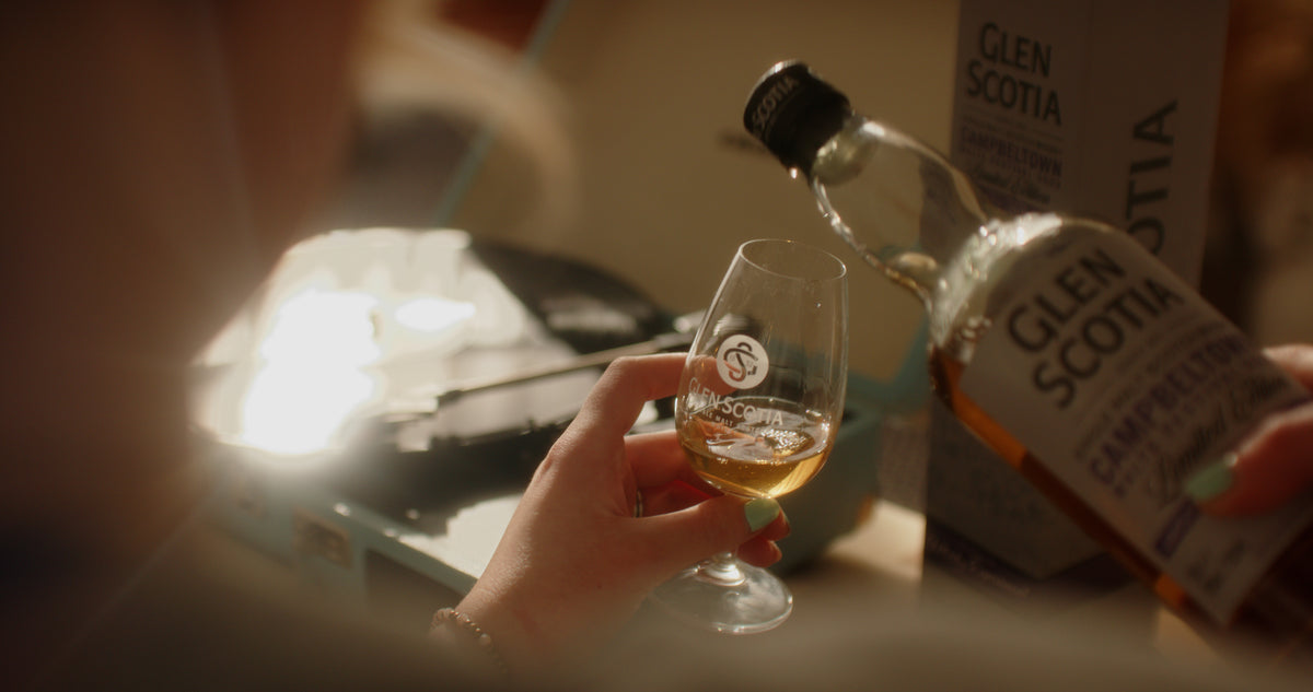 A person is holding a glass with a small amount of amber-colored The Sound of Glen Scotia whisky while another hand pours more from the bottle into the glass. Both the bottle and the glass proudly display the Glen Scotia logo, capturing a perfect Campbeltown moment against a soft, blurred background.