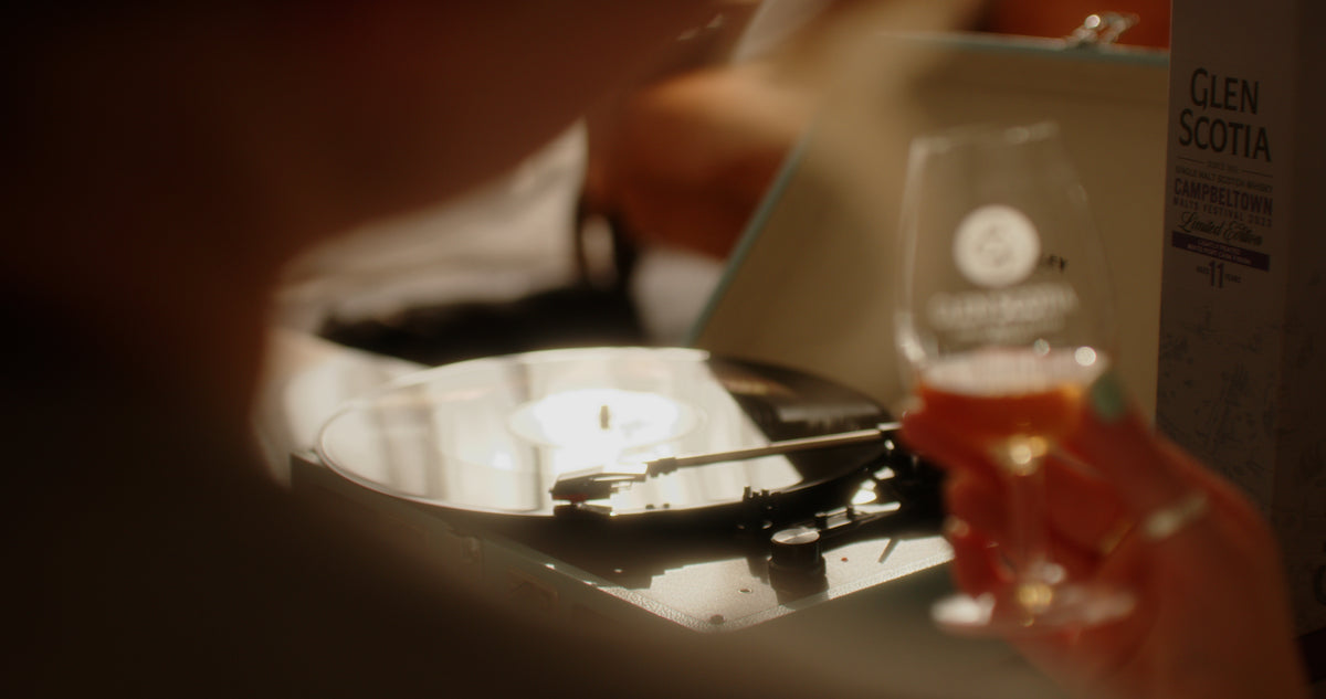 A close-up of a vinyl record playing on a turntable. In the foreground to the right, a person holds a glass with some amber-colored The Sound of Glen Scotia whisky. The glass has a blurred Glen Scotia logo on it. The scene is illuminated with warm lighting, giving it a cozy ambiance.