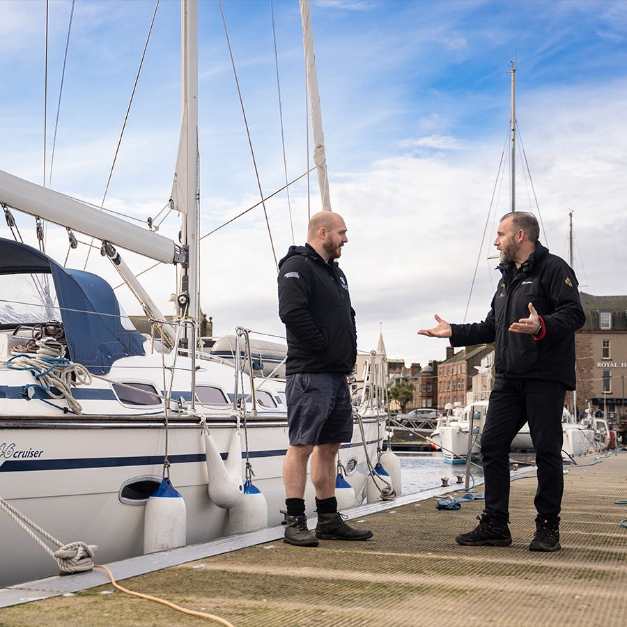 Two men stand on a dock beside a sailboat in a marina, engaged in conversation. One man is dressed in a black jacket and shorts, the other in a black jacket and pants. The background features other boats and buildings against a partly cloudy sky.