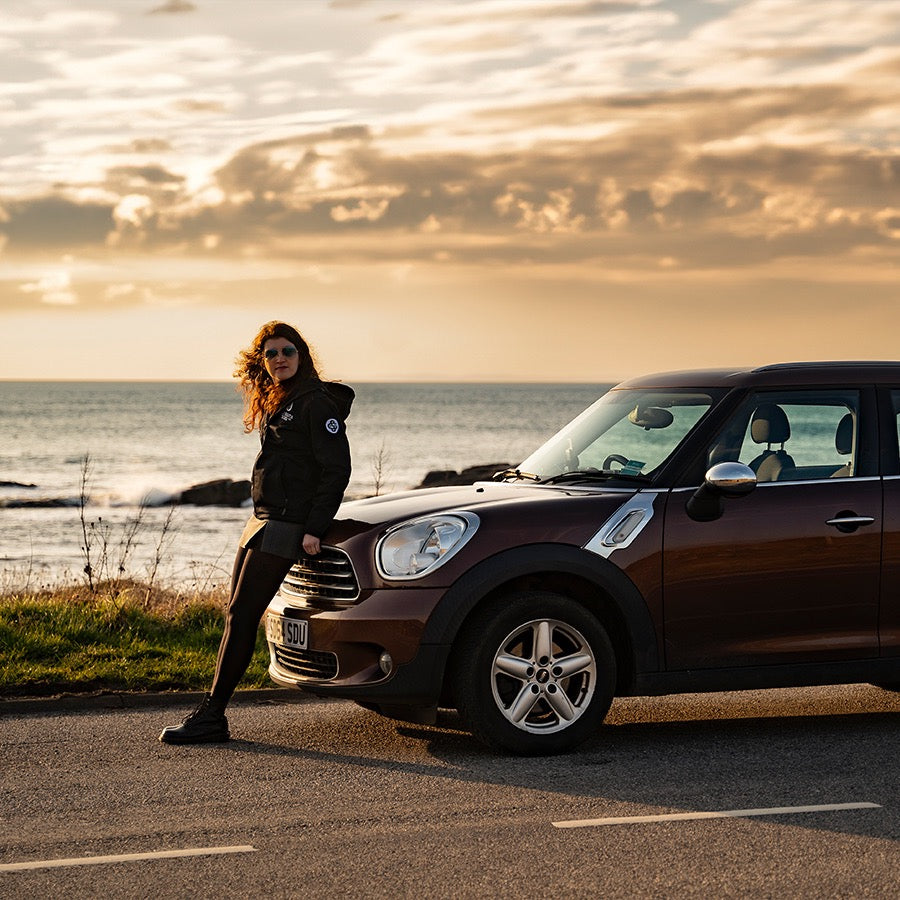 A woman with wavy hair in a dark jacket and sunglasses leans against a brown Mini Cooper parked on a coastal road. The sun is low in the sky, casting a golden light over the ocean and the clouds.