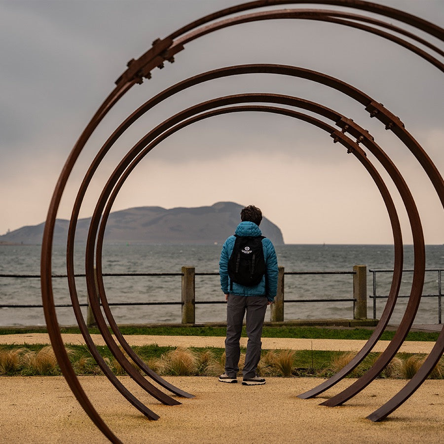 A person wearing a blue jacket and a backpack stands on a sandy path between large metal rings, overlooking a cloudy seascape with an island in the background. The scene is framed by tall grass and a fence along the waterfront.