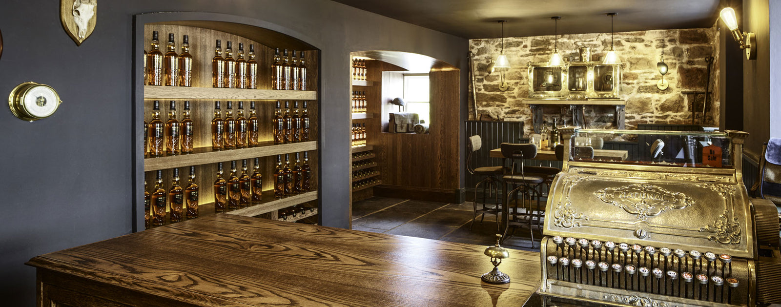 A cozy, rustic store or bar with stone walls and wooden shelving stocked with numerous bottles of alcohol. A vintage brass cash register sits on a wooden counter in the foreground. Warm lighting and a small window create an inviting ambiance.