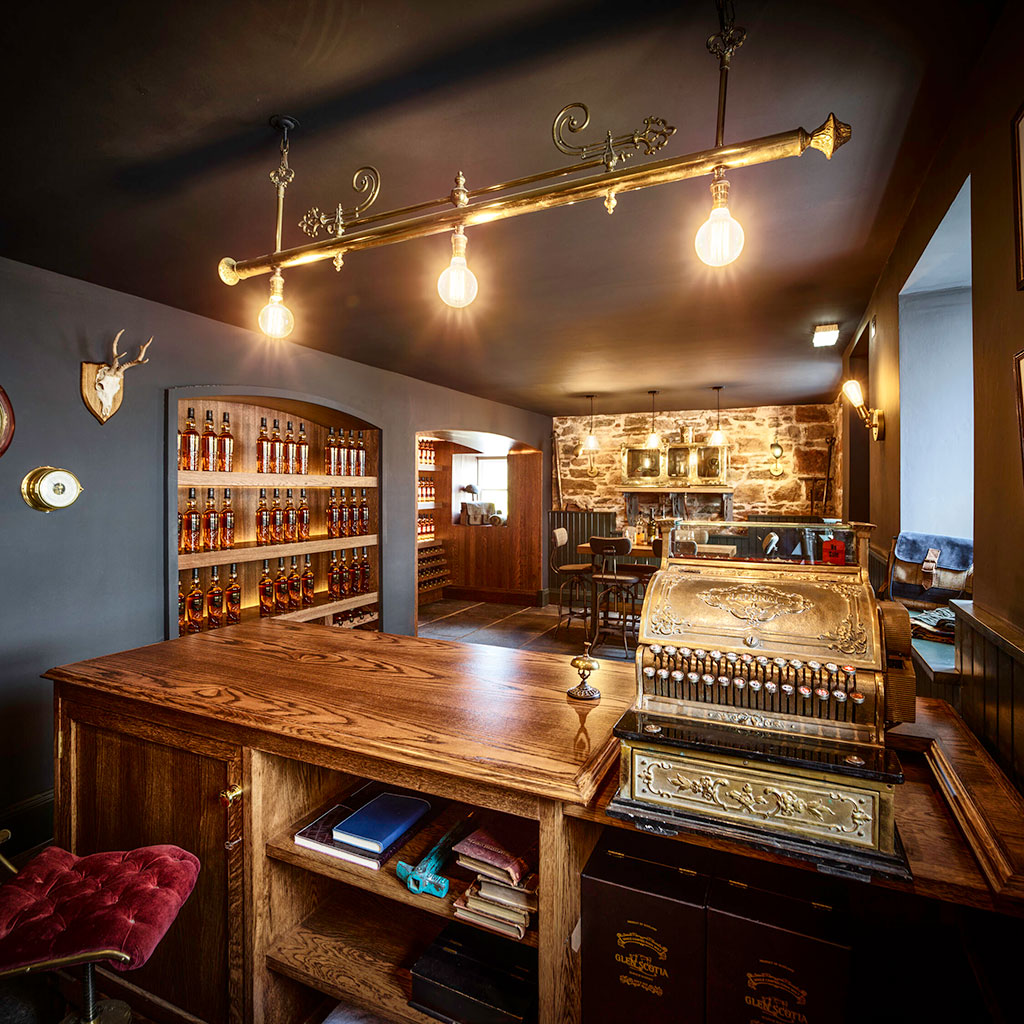 A vintage-themed bar features a wooden counter with an ornate cash register, shelves stocked with bottles, exposed brick walls, and warm lighting from hanging Edison bulbs. The atmosphere exudes a rustic charm, enhanced by various decorative accents.