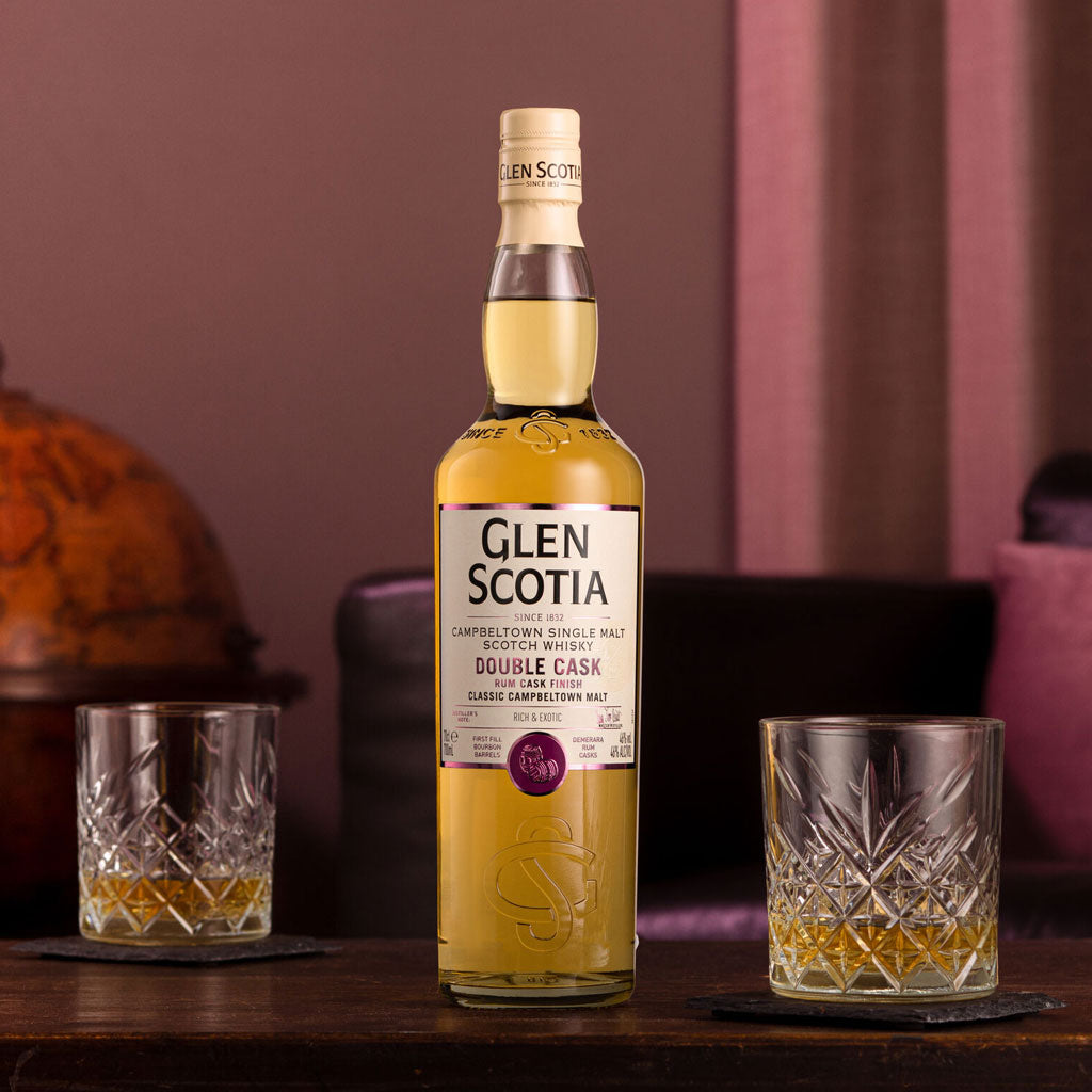 A bottle of Glen Scotia Double Cask single malt Scotch whisky stands on a dark wooden table with two crystal glasses of whisky placed on either side. The background features soft, warm lighting with a globe and draped fabric creating a cozy ambiance.