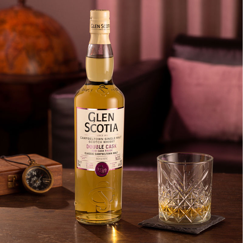 A bottle of Glen Scotia double cask Scotch whisky sits on a wooden table next to a filled crystal glass. An old-fashioned pocket watch and a globe are in the blurred background. The setting appears warm and cozy, with dim lighting.