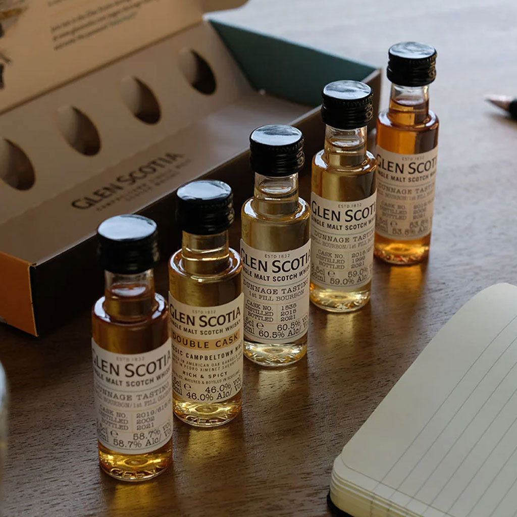 Five small bottles of Glen Scotia single malt Scotch whiskey are lined up on a wooden table. Each bottle has a label detailing its type and alcohol content. An open box, originally containing the bottles, and a notebook are visible in the background.