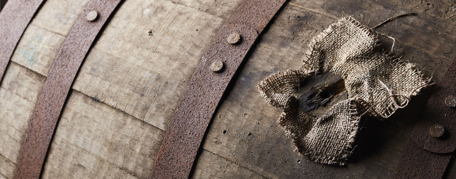 A close-up shot of an old wooden barrel secured with metal straps. A small, beige burlap patch is affixed to the barrel's surface, hinting at a makeshift repair. The wood's texture and metal straps appear weathered, showcasing signs of age and use.