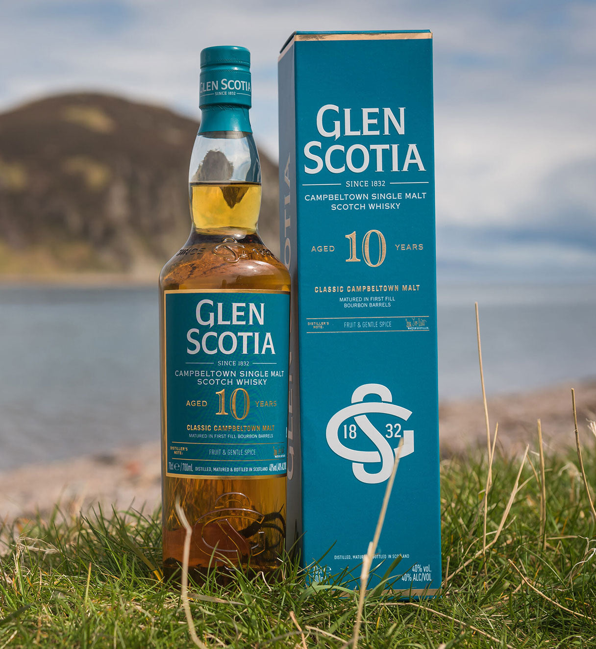 A bottle of Glen Scotia 10-year-old Campbeltown single malt Scotch whisky stands next to its teal box on grass, with a scenic backdrop of a hill and water under a cloudy sky. The box and bottle are labeled with details including "Established 1832" and "Classic Campbeltown Malt.