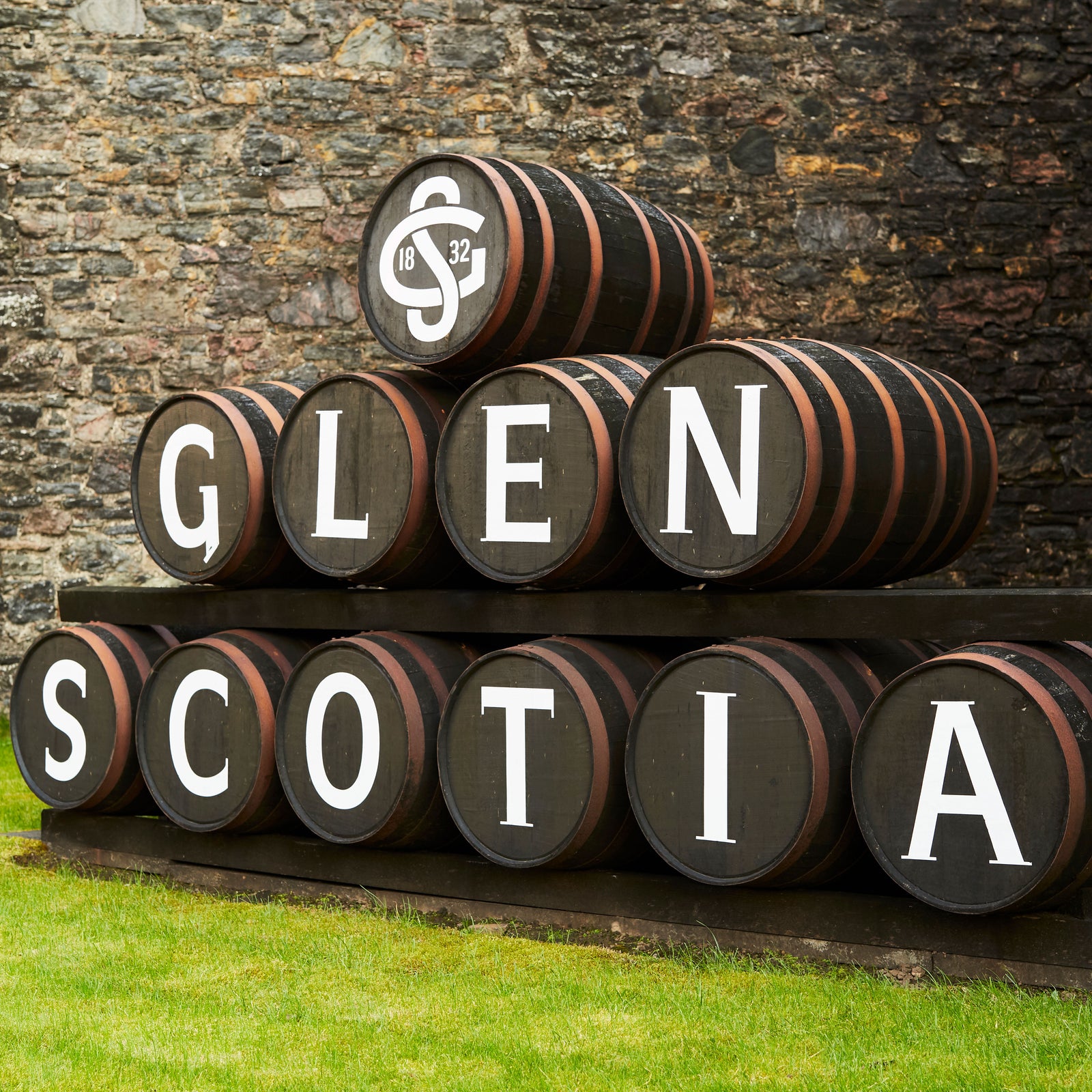 Stacked wooden barrels with "GLEN SCOTIA" written on them in bold white letters. The background features a stone wall and green grass in the foreground.