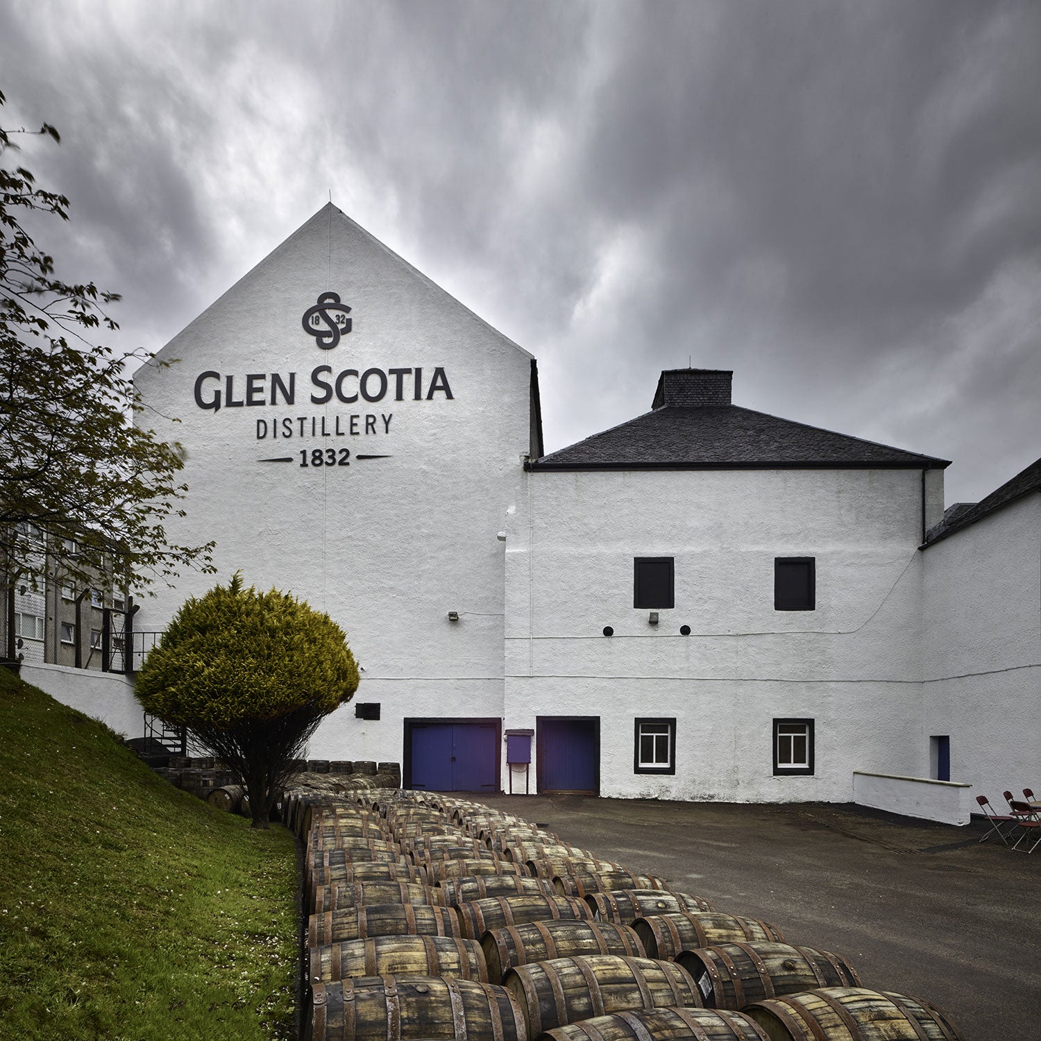 The exterior of the Glen Scotia Distillery, a white building with the distillery's name and 1832 on the front. Barrels are stacked in front of the building. The sky is overcast. A green bush is on the left side of the image.