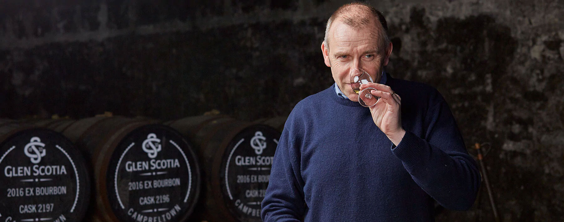 A man in a blue sweater smells a glass of whisky. Behind him are several barrels labeled "Glen Scotia 2015 Ex Bourbon Cask 2197," stacked against a dark stone wall. The setting appears to be a distillery.