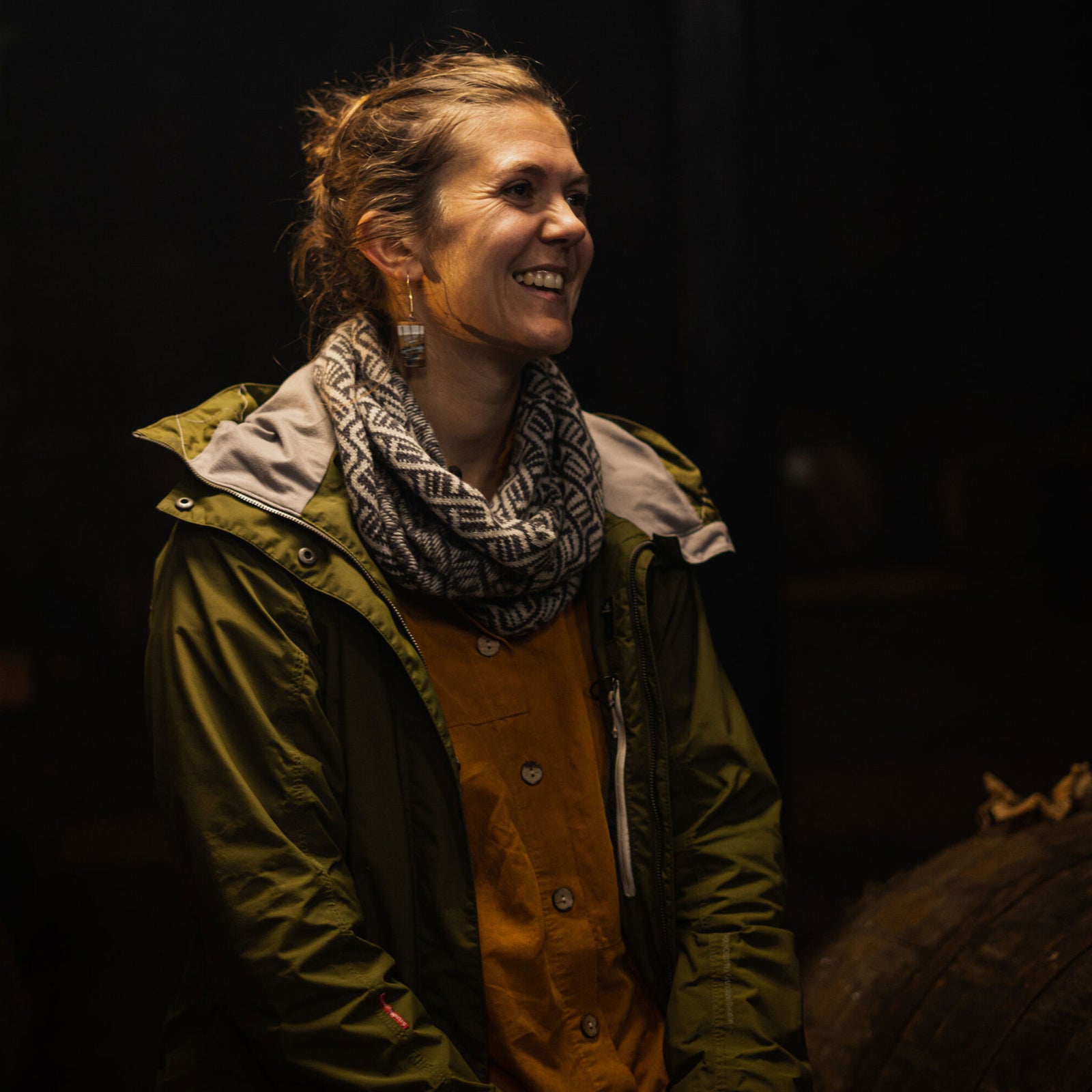 A woman with a smile stands indoors wearing a green jacket over a mustard-colored shirt and a patterned scarf. The background is dimly lit and blurry, with a dark ambiance creating a cozy atmosphere.
