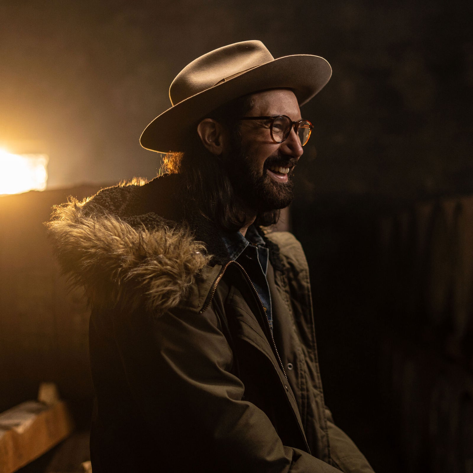 A person with long hair, glasses, and a beard is seen smiling and laughing. They are wearing a wide-brimmed hat and a coat with a fur-trimmed hood. The background is dimly lit with a warm light source creating a cozy atmosphere.