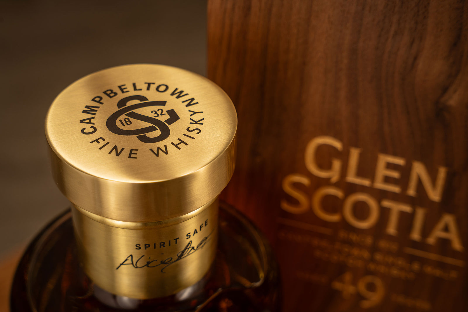 Close-up of a bottle of Campbeltown Fine Whisky with a golden cap labeled "Spirit Safe." Beside the bottle is a wooden box inscribed with "Glen Scotia" and "49," indicating the age of the whisky. The image highlights the luxurious packaging and branding.