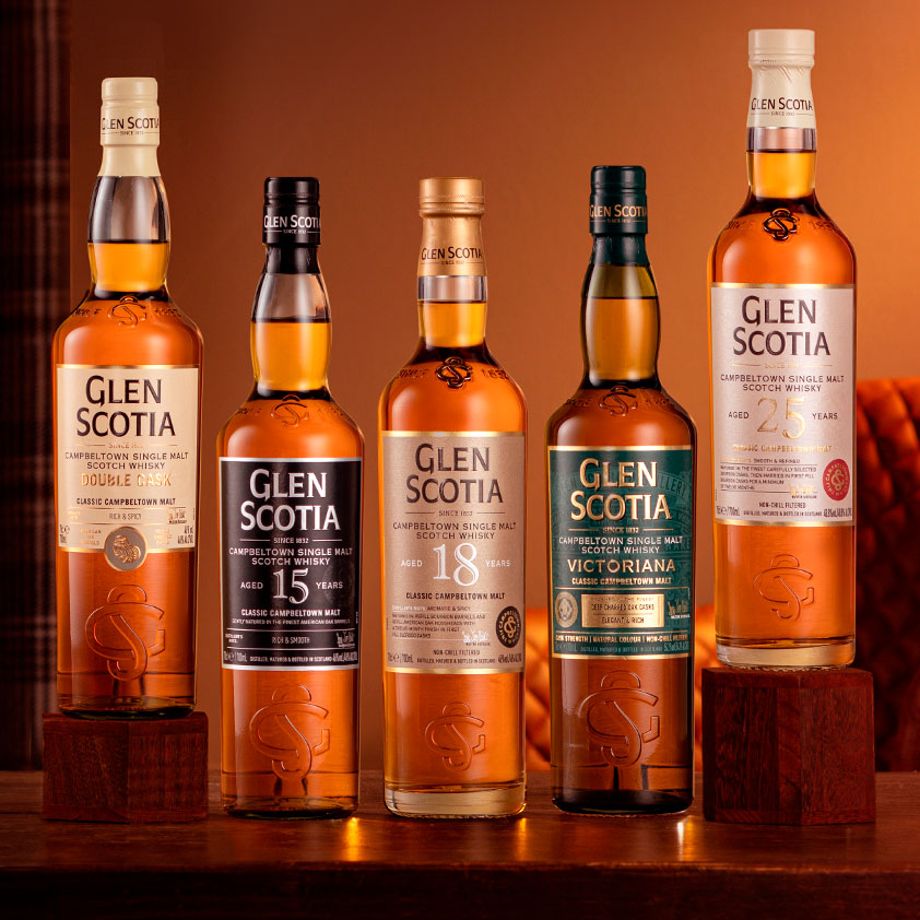 A lineup of five Glen Scotia whisky bottles. From left to right: Double Cask, 15 Years Old, 18 Years Old, Victoriana, and 25 Years Old. They are displayed against a warm-toned background on wooden platforms.