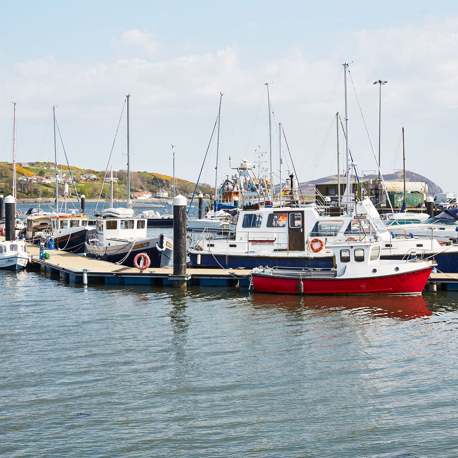 A small marina with several boats docked at the pier. The boats vary in size and color, with a prominent red and white boat in the foreground. The water is calm and the background features a hilly landscape under a partly cloudy sky.