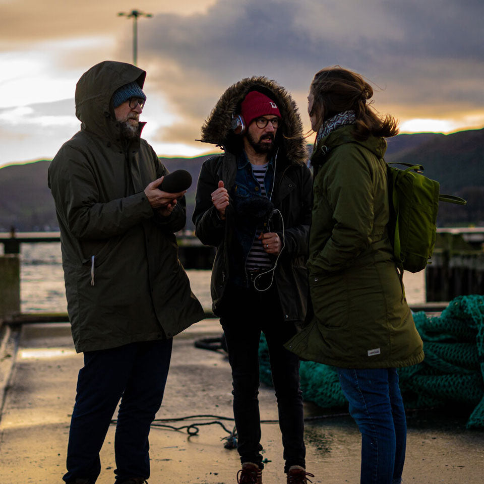 Three individuals stand on a dock having a conversation at dusk. Two wear green jackets and the third person, in the middle, wears a red beanie and headphones, holding a microphone. The background shows water, mountains, and a cloudy sky.