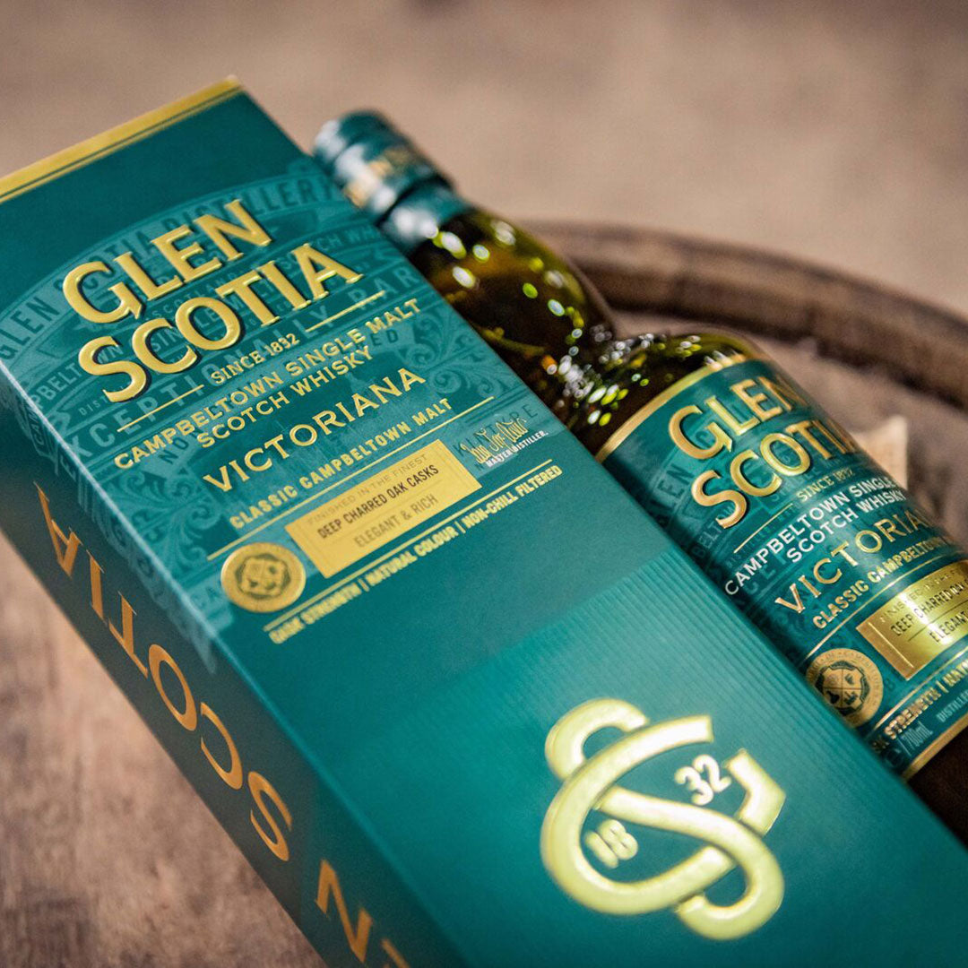 A Glen Scotia box and bottle are displayed. The teal packaging features gold text, highlighting "Campbeltown Single Malt Scotch Whisky," "Victoriana," and their 1832 heritage. The bottle lies alongside the box, both resting on a wooden surface.