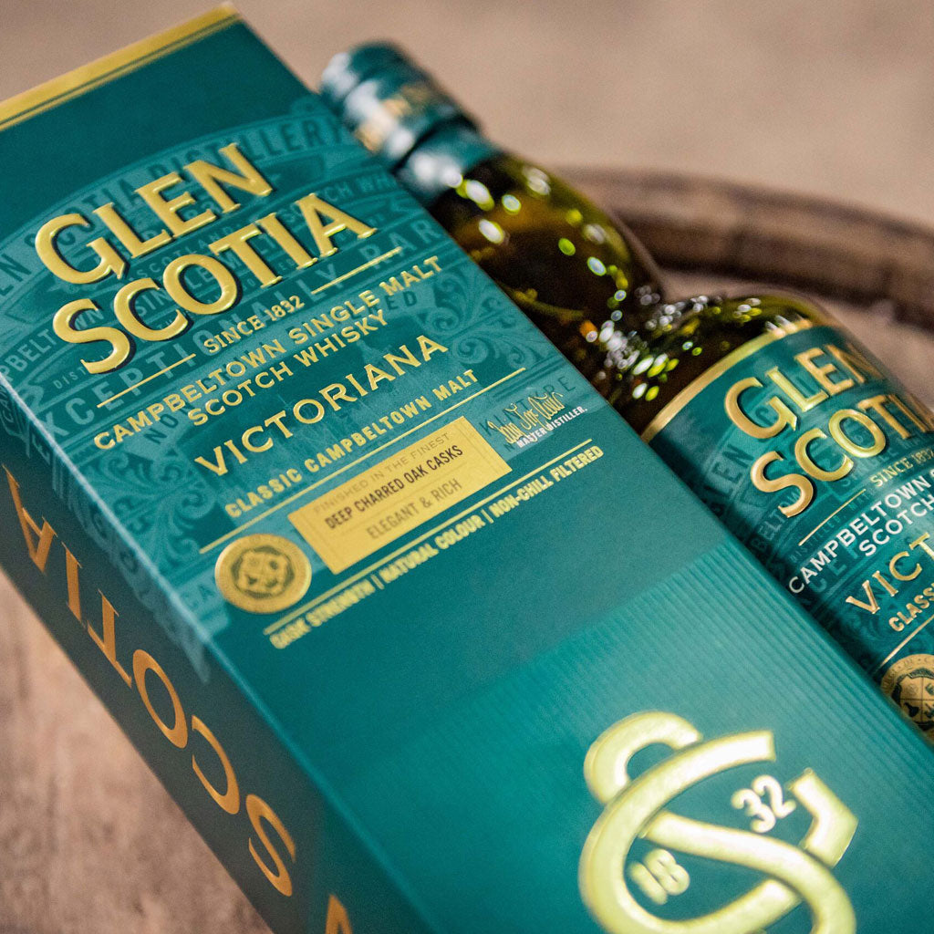 A green and gold box of Glen Scotia Victoriana Campbeltown single malt Scotch whisky is displayed with the bottle partially out of the box. The packaging highlights its classic Campbeltown malt and elegant finish, emphasizing its classic cask origins.